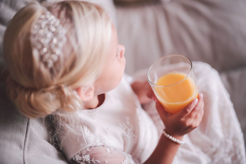 Person holding orange juice glass wearing white lace dress with elegant updo hairstyle.
