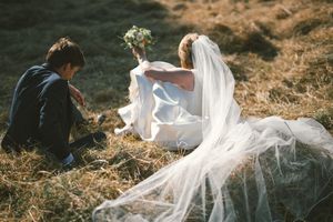 A romantic wedding scene on a sunlit hillside with a flowing white veil and dress against dried grass.
