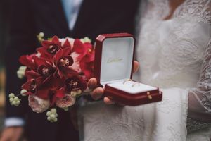 A red velvet ring box and vibrant red bouquet held against a white wedding dress background.