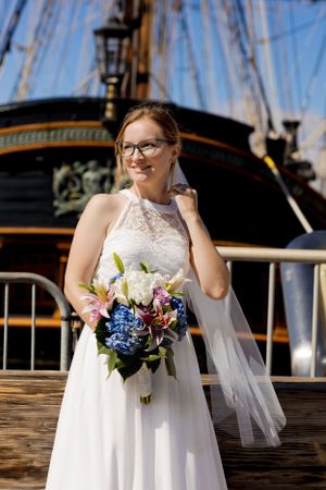 A bride in a white lace dress holds a blue and white bouquet on a wooden deck near a ship.Shot By Zeus Holbrook San Diego Wedding Photograph