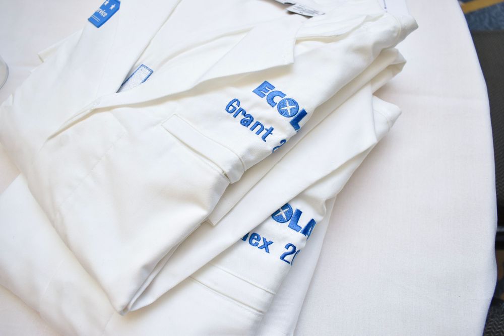 Folded white garments with blue ECOL Grant text printed on them, stacked neatly on a white surface.