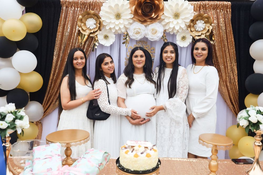 Group in white outfits poses at baby shower with gold backdrop, balloons, and floral decorations.