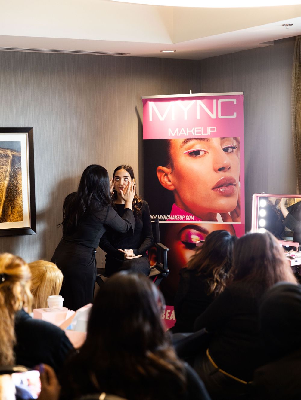 MYNC Makeup artist applying cosmetics on model at a beauty demonstration event with audience watching.