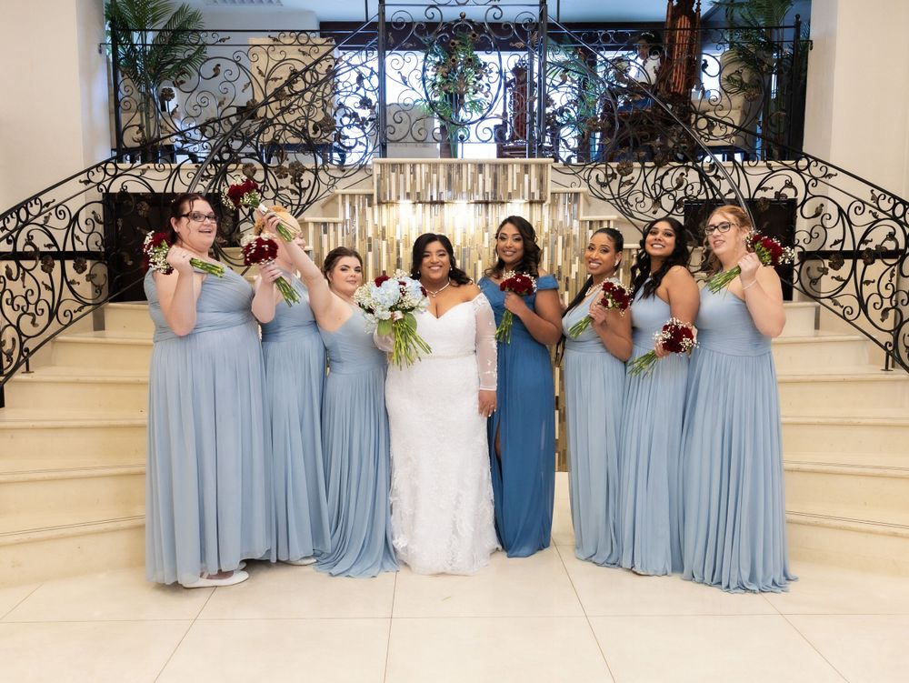 Wedding party poses on elegant white staircase with ornate railings in light dusty blue gowns.
