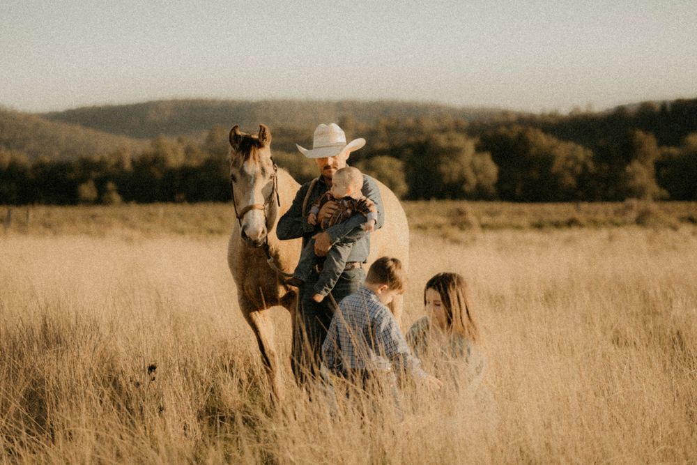 A family in western attire stands together in a golden field at sunset with their horse behind rolling hills.