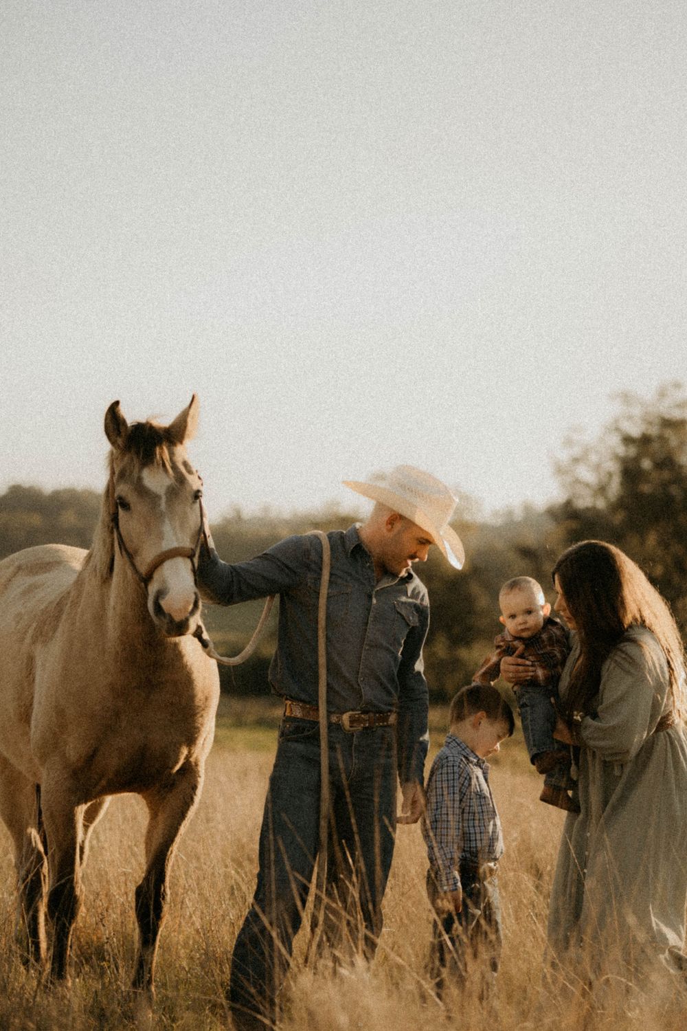 A family stands together in a golden field at sunset with their dun-colored horse.