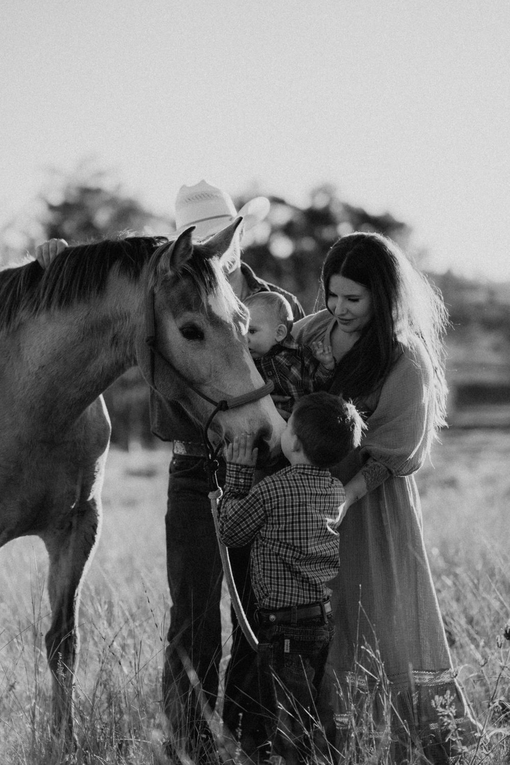 A mother and child stand together in a field petting a horse in a dreamy black and white photograph.