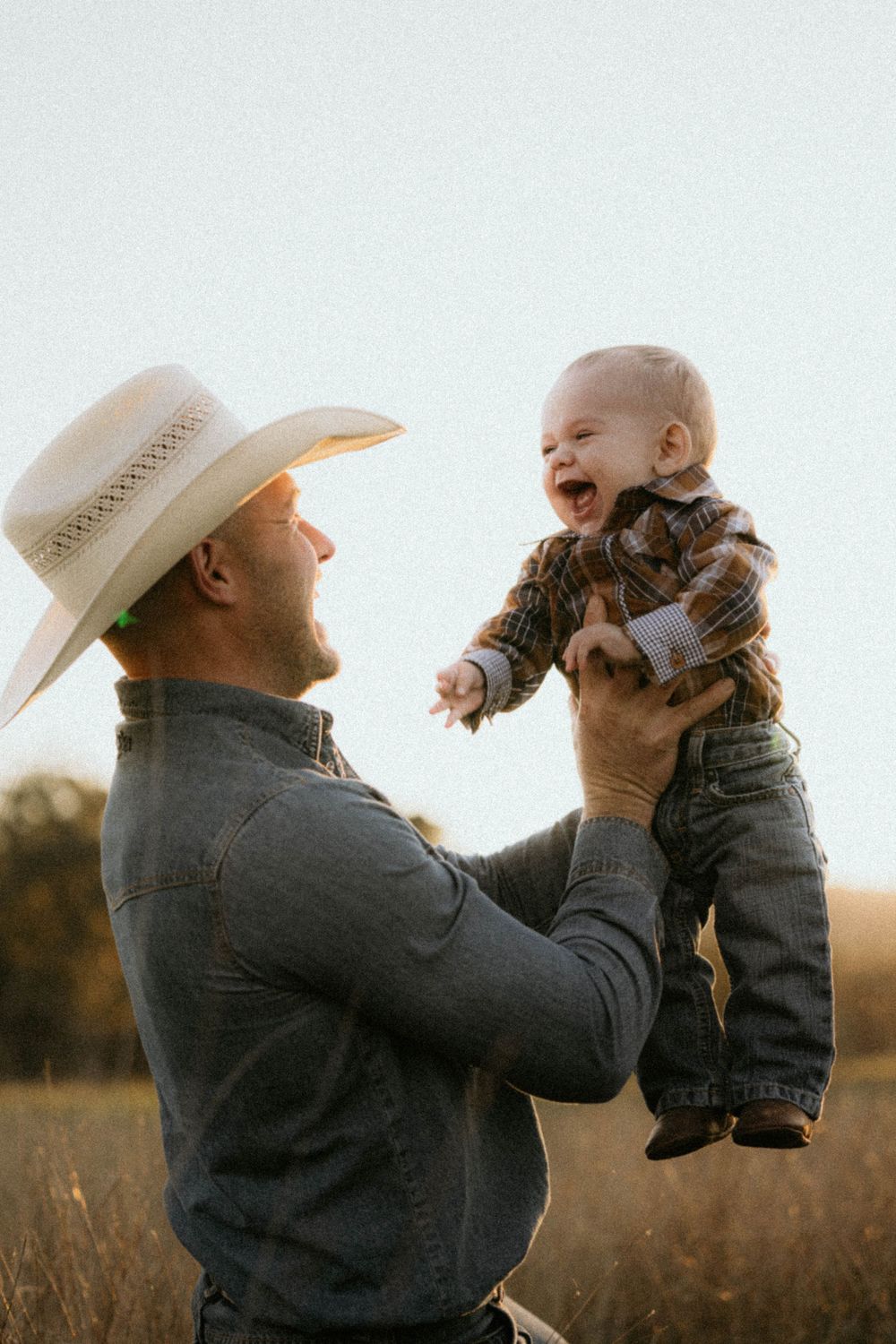 A person wearing a cowboy hat lifts a laughing baby dressed in denim overalls in a sunlit field at dusk.