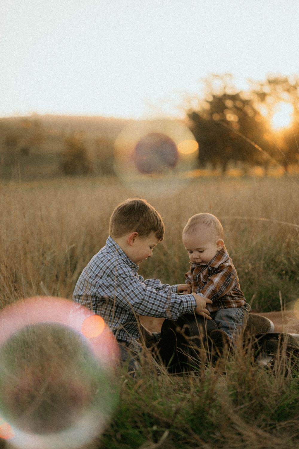 Two children sit together in a grassy field during golden hour sunset with lens flares.