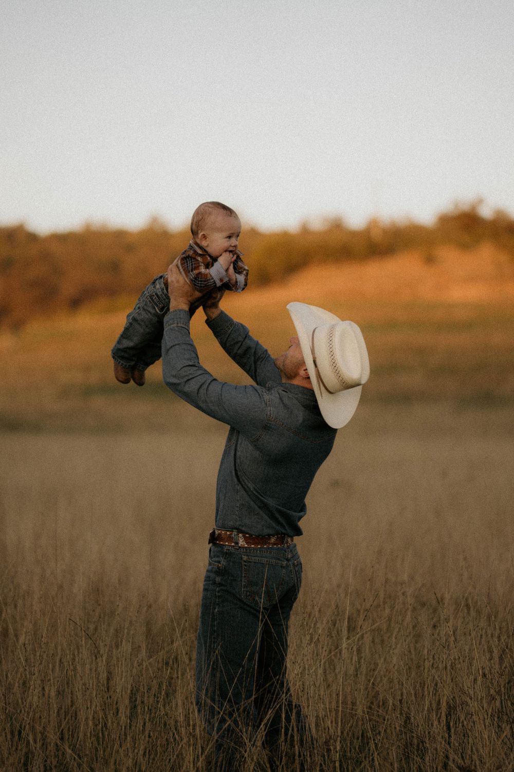A person in a cowboy hat lifts a small baby into the evening sky in a golden field at sunset.