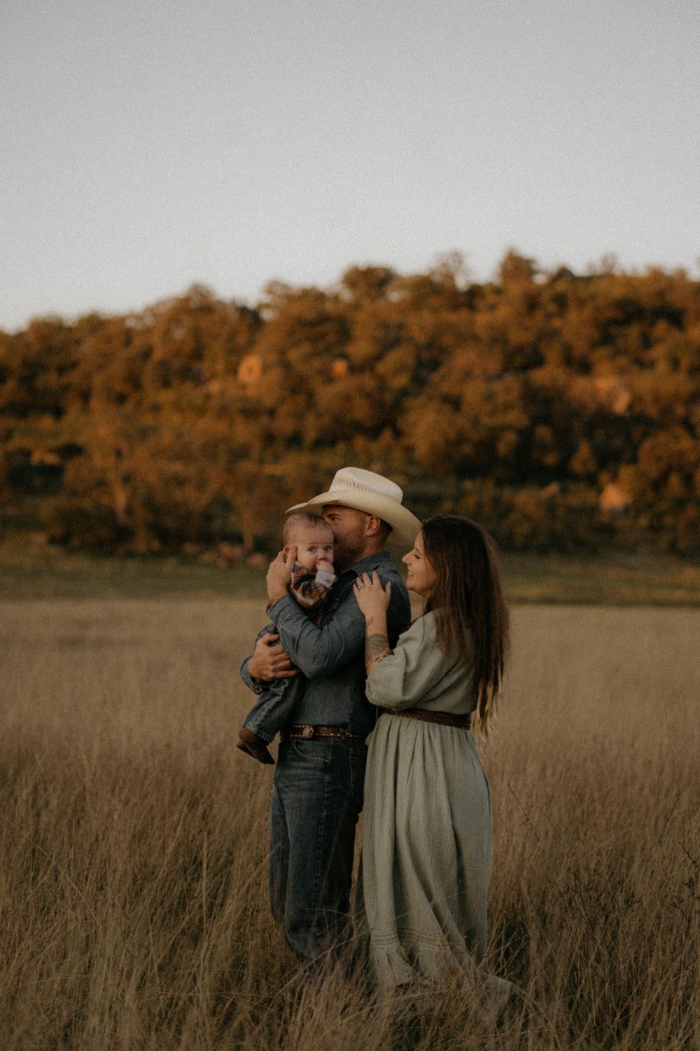 Country family stands together in a golden autumn field at sunset with warm fall colors in the background.