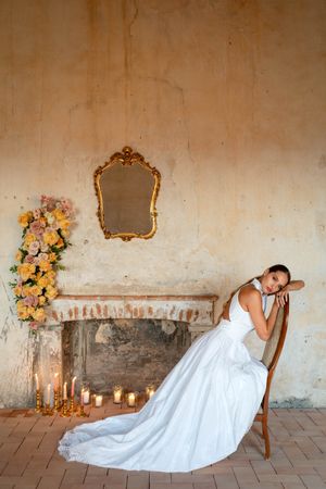 Elegant bridal scene with white gown against rustic wall with floral arrangements and candles.