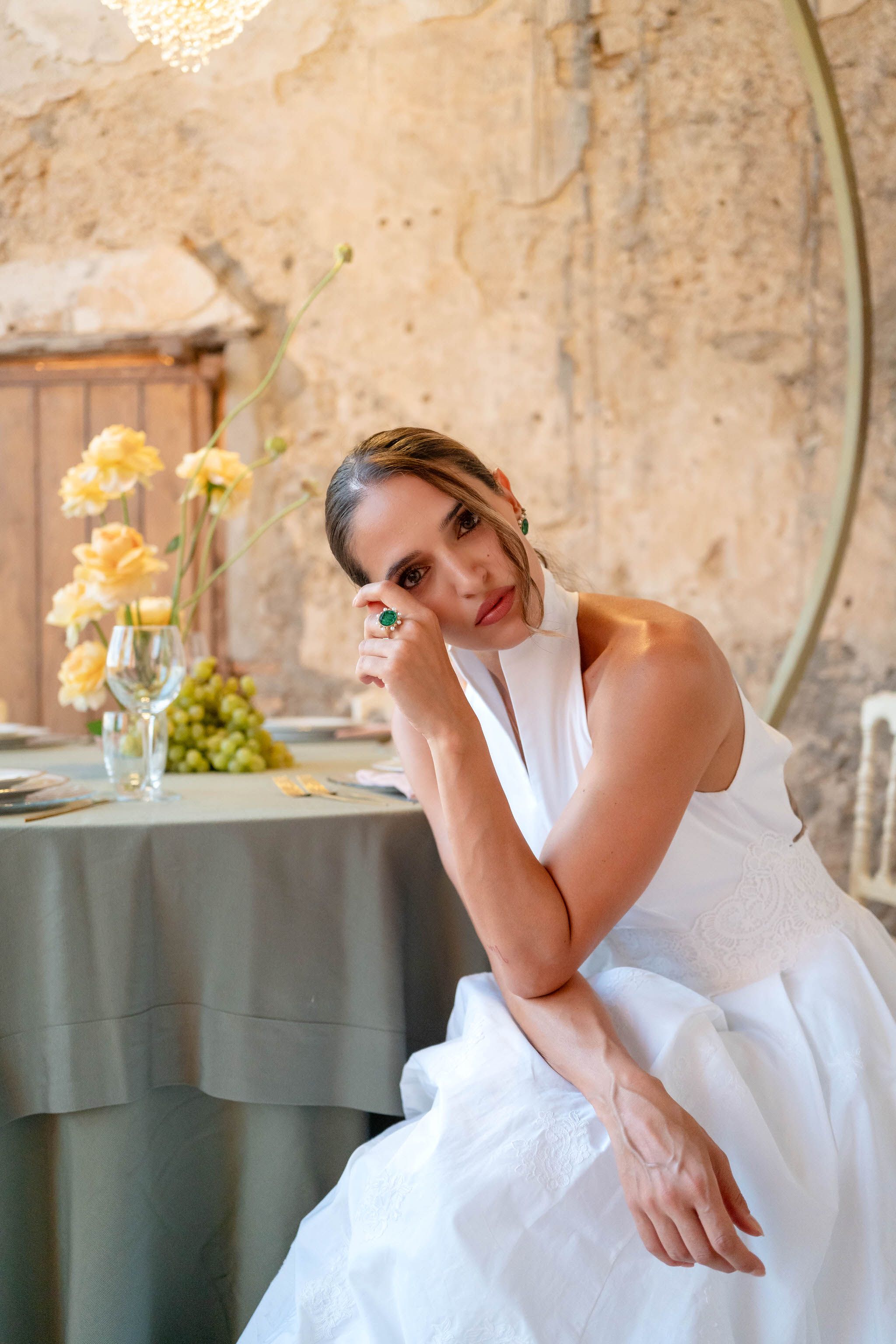 A bride in a white wedding dress leans against a table at a reception venue with stone walls and floral centerpieces.
