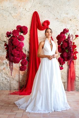 A figure in a flowing white lace dress poses elegantly with vibrant red fabric and deep crimson flowers against a rustic wall.