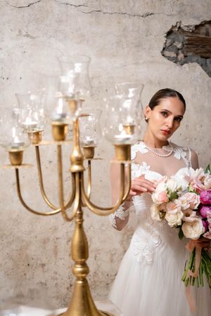 Elegant bridal portrait with gold candelabra and mixed pastel bouquet against rustic wall.