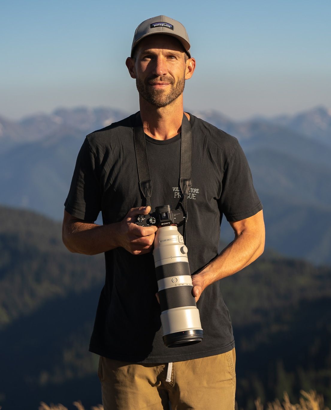 Photographer wearing black shirt and cap holds professional camera lens against mountain landscape backdrop during golden hour.