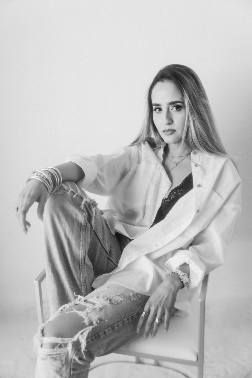 Fashion model in white shirt and distressed jeans poses on chair in minimalist studio setting.