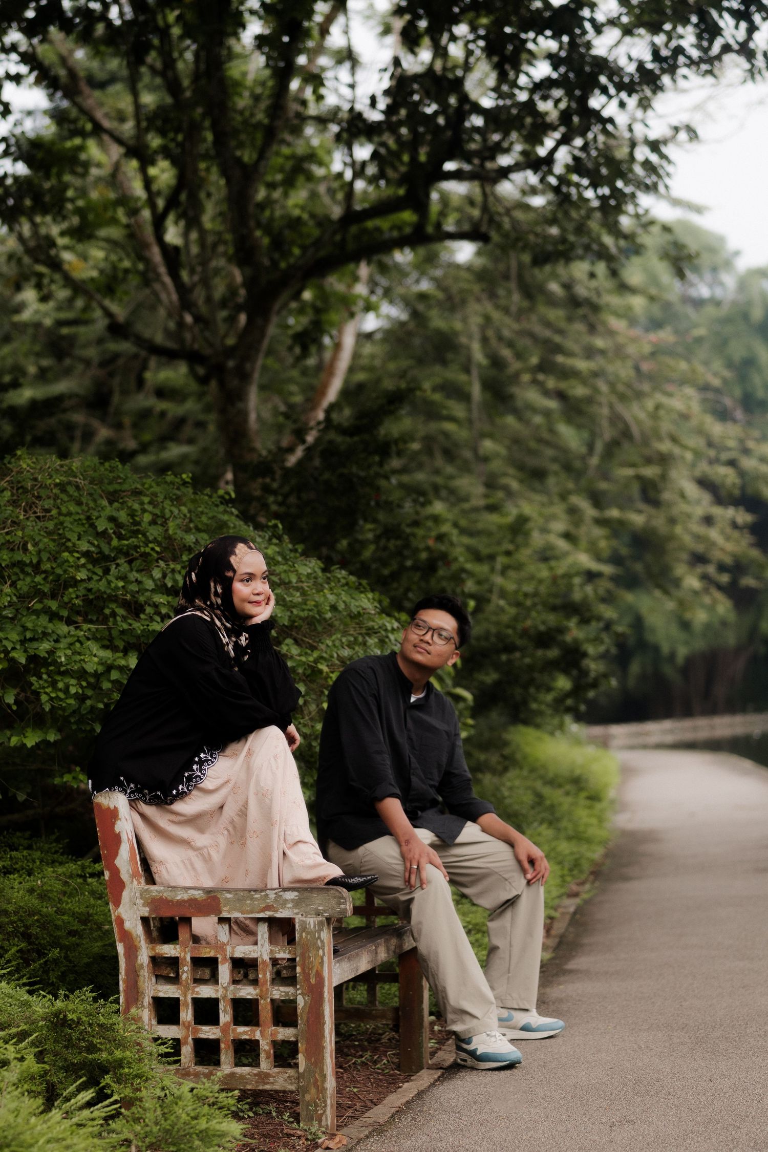 A couple sits on a wooden bench in a lush green park, woman in hijab and black top, man in black shirt.
