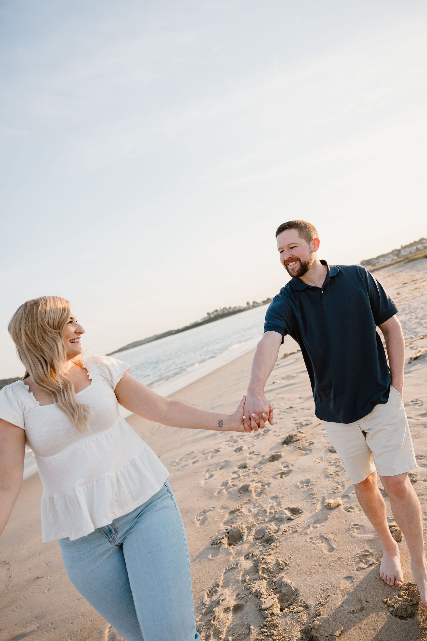 A smiling couple holds hands while walking on a sandy beach at sunset, woman in white top and jeans, man in navy shirt.
