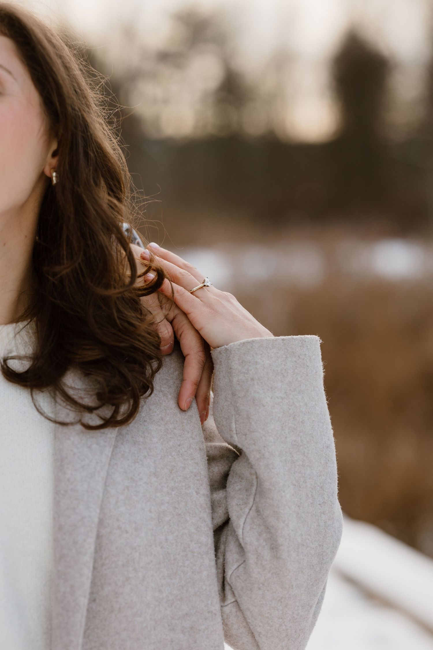 A woman in a gray coat rests her hand on her shoulder, wearing a gold ring, with a snowy outdoor backdrop.