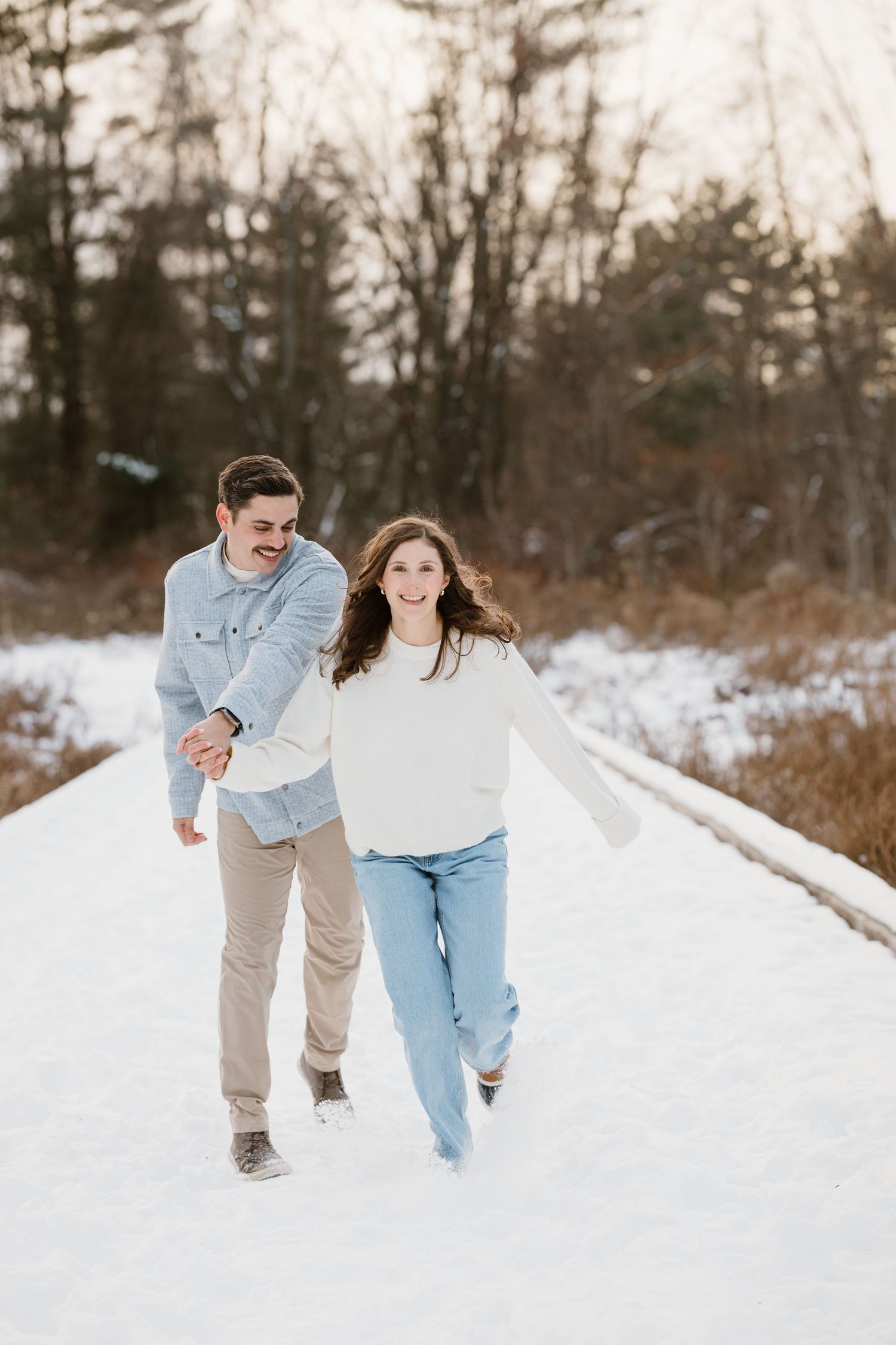 A happy couple holds hands and runs together on a snowy path surrounded by winter trees in soft golden light.