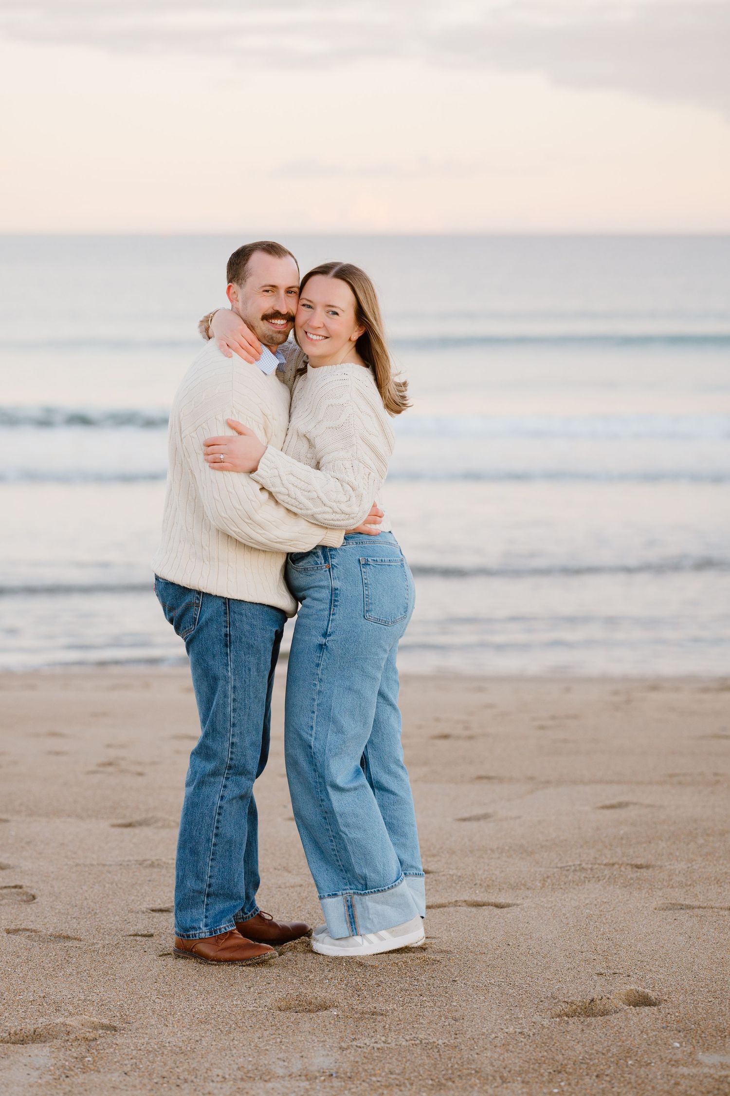 A couple embracing on a sandy beach at sunset, both wearing cream sweaters and jeans, smiling warmly.