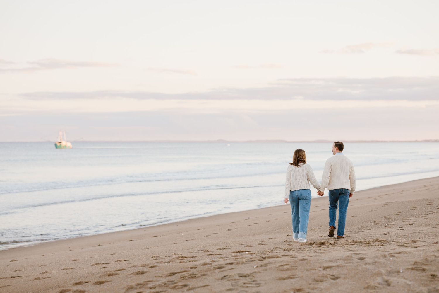 Couple holding hands walking on a sandy beach at sunset with a ship visible on the calm ocean horizon.