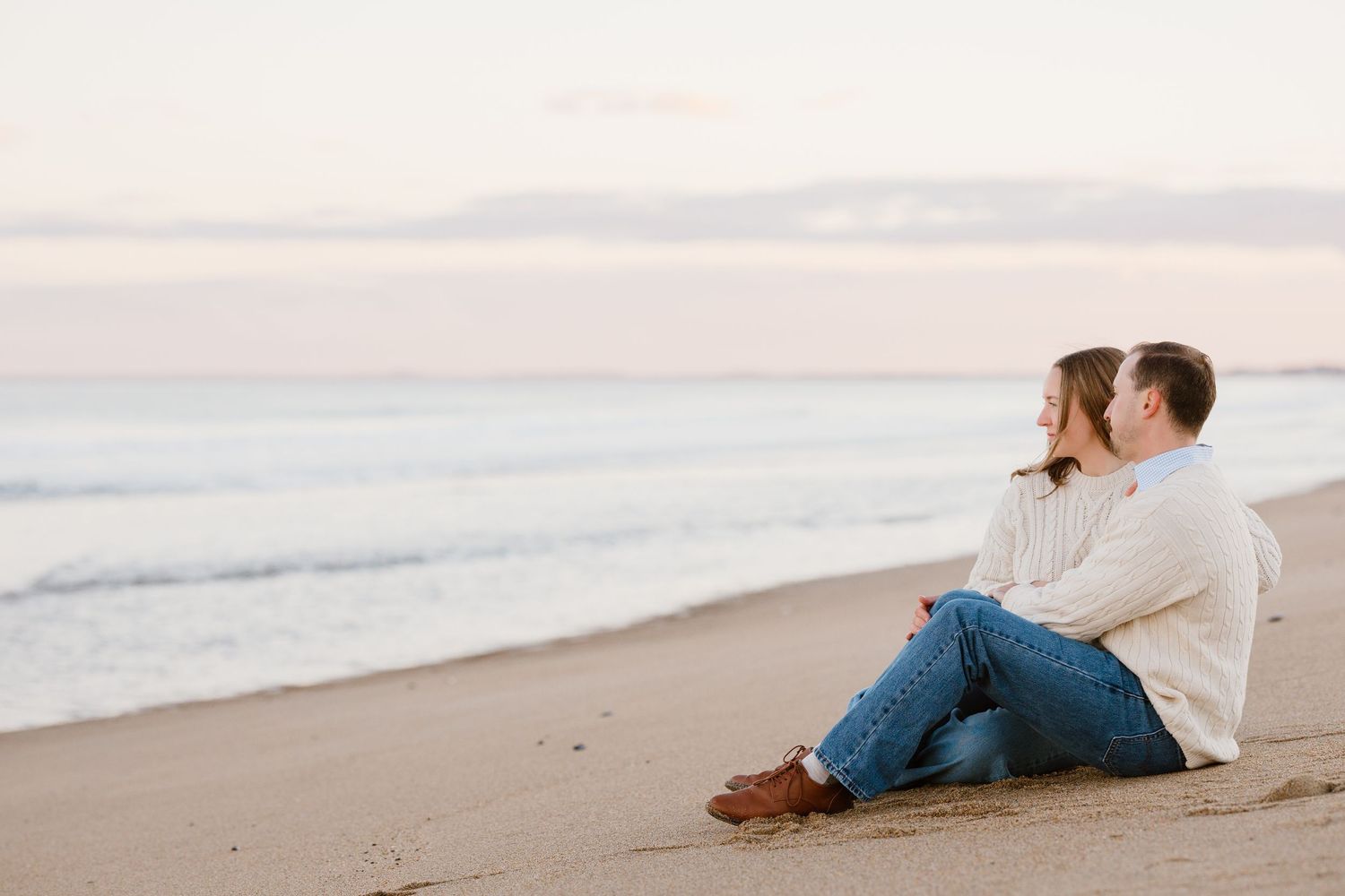 Couple sitting together on a sandy beach at sunset, gazing at calm ocean waves in a romantic seaside moment.