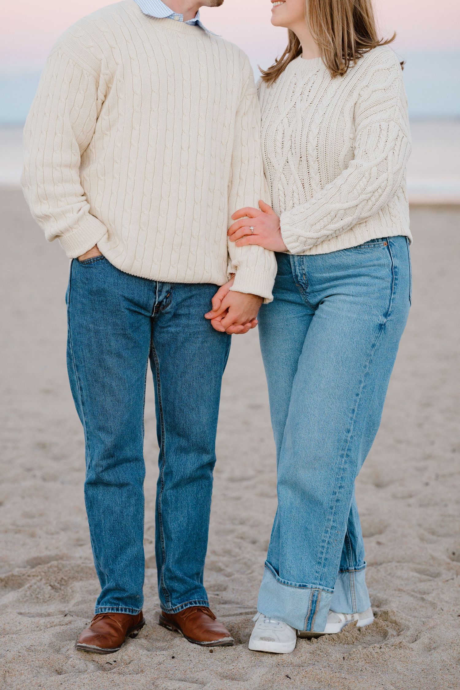 Couple in matching cream cable-knit sweaters and blue jeans holding hands on a sandy beach at dusk.