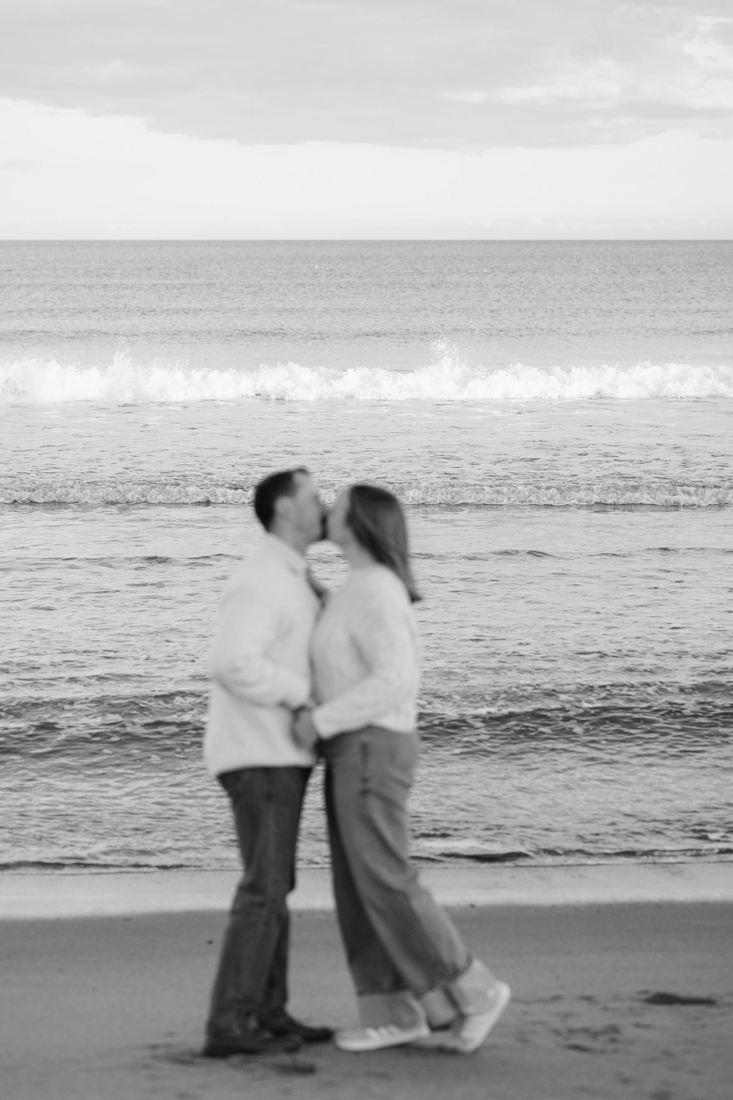 Black and white photo of a couple kissing on a beach with ocean waves in the background.