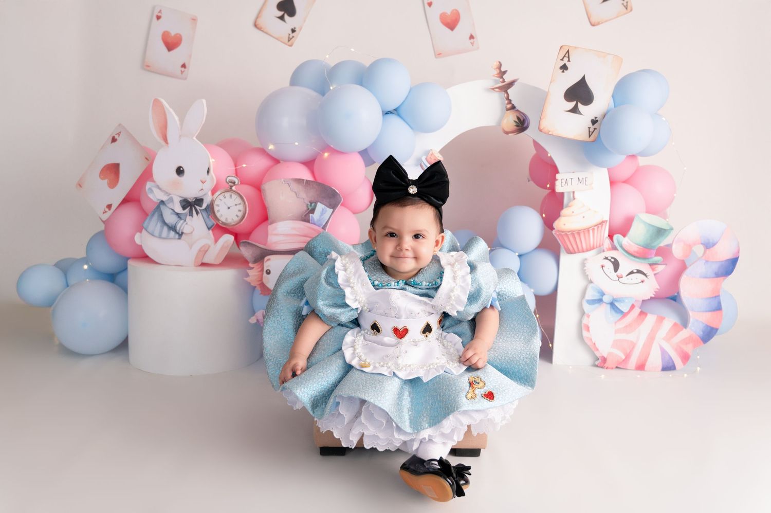 Alice in Wonderland themed baby photoshoot with blue balloons and white rabbit decorations.