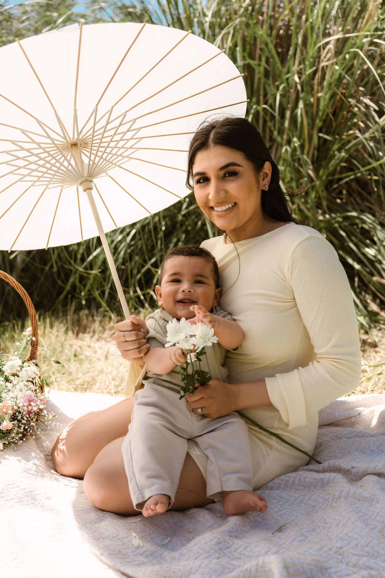 Mother and child lounging on blanket with white parasol in sunny outdoor setting.