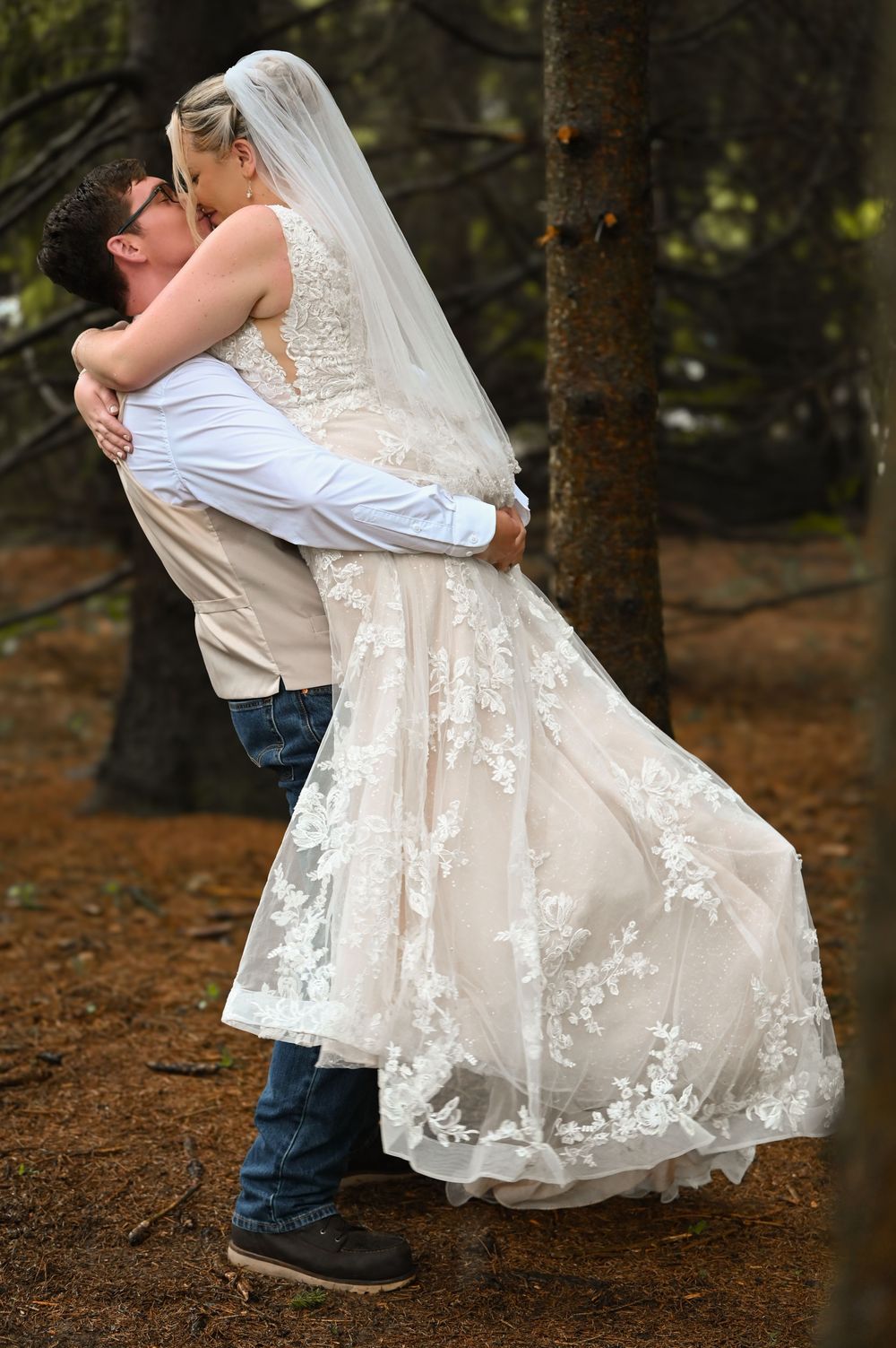 Groom lifting bride in lace wedding dress for a romantic kiss among tall trees in a forest.