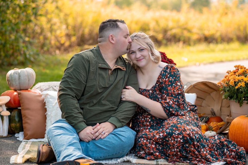 Couple sits together on blanket surrounded by pumpkins and mums during autumn photo session.