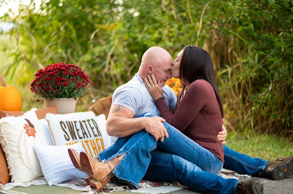 Couple shares a kiss during fall-themed outdoor picnic session with pumpkins and autumn decor.
