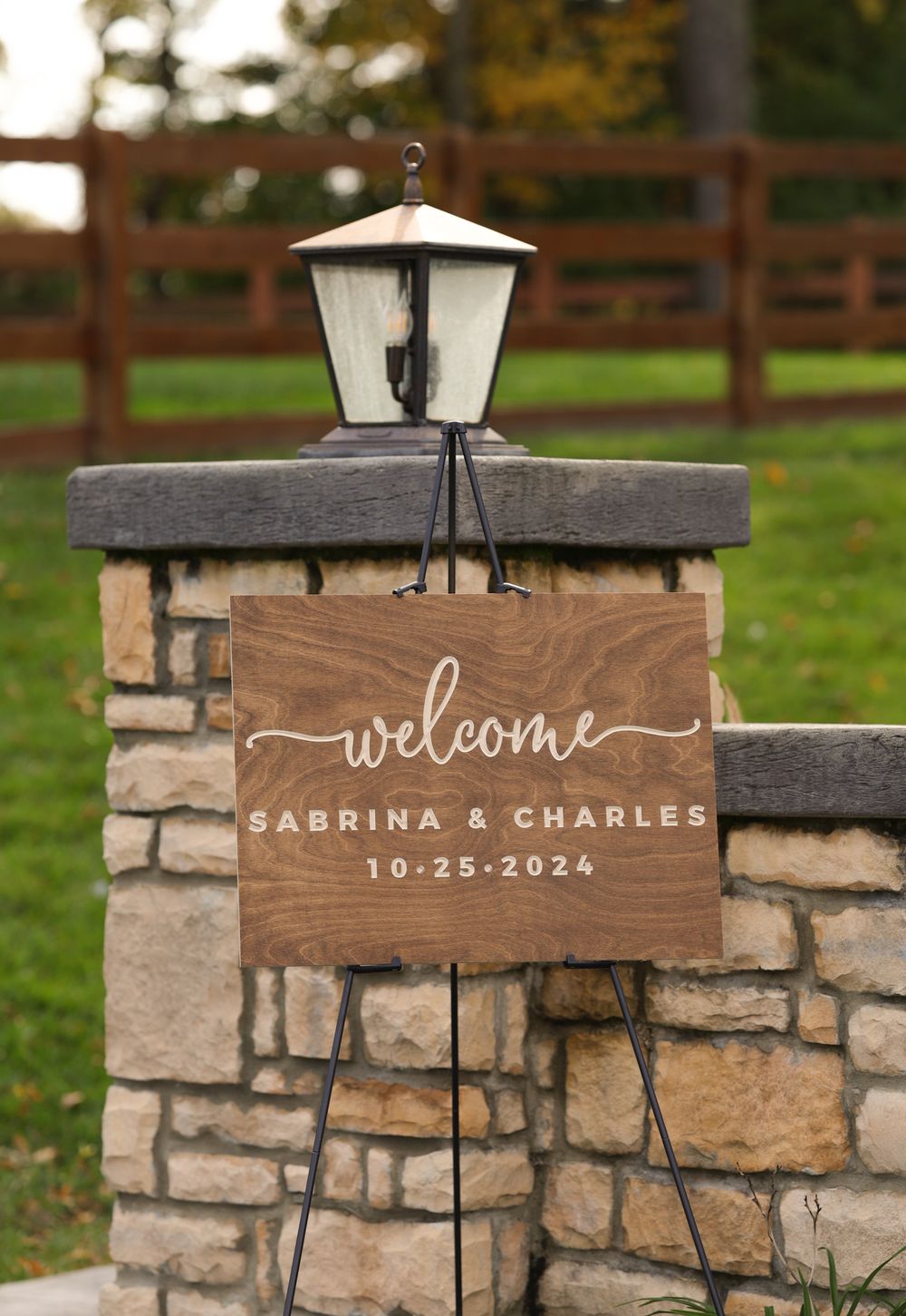 Wooden welcome sign for Sabrina and Charles wedding dated 10-25-2024 on a stone pillar outdoors.