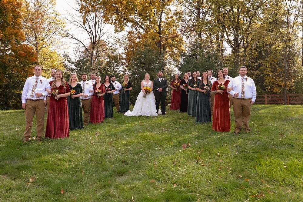 Large wedding party in burgundy and green dresses with groomsmen posing on a green lawn in fall.