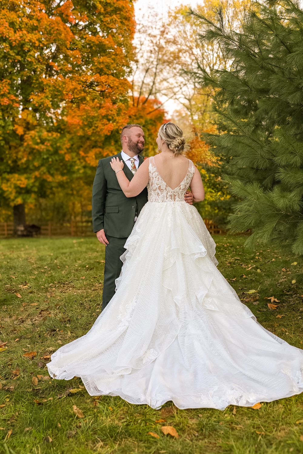 Bride in layered white gown and groom in green suit sharing a moment among vibrant autumn foliage.