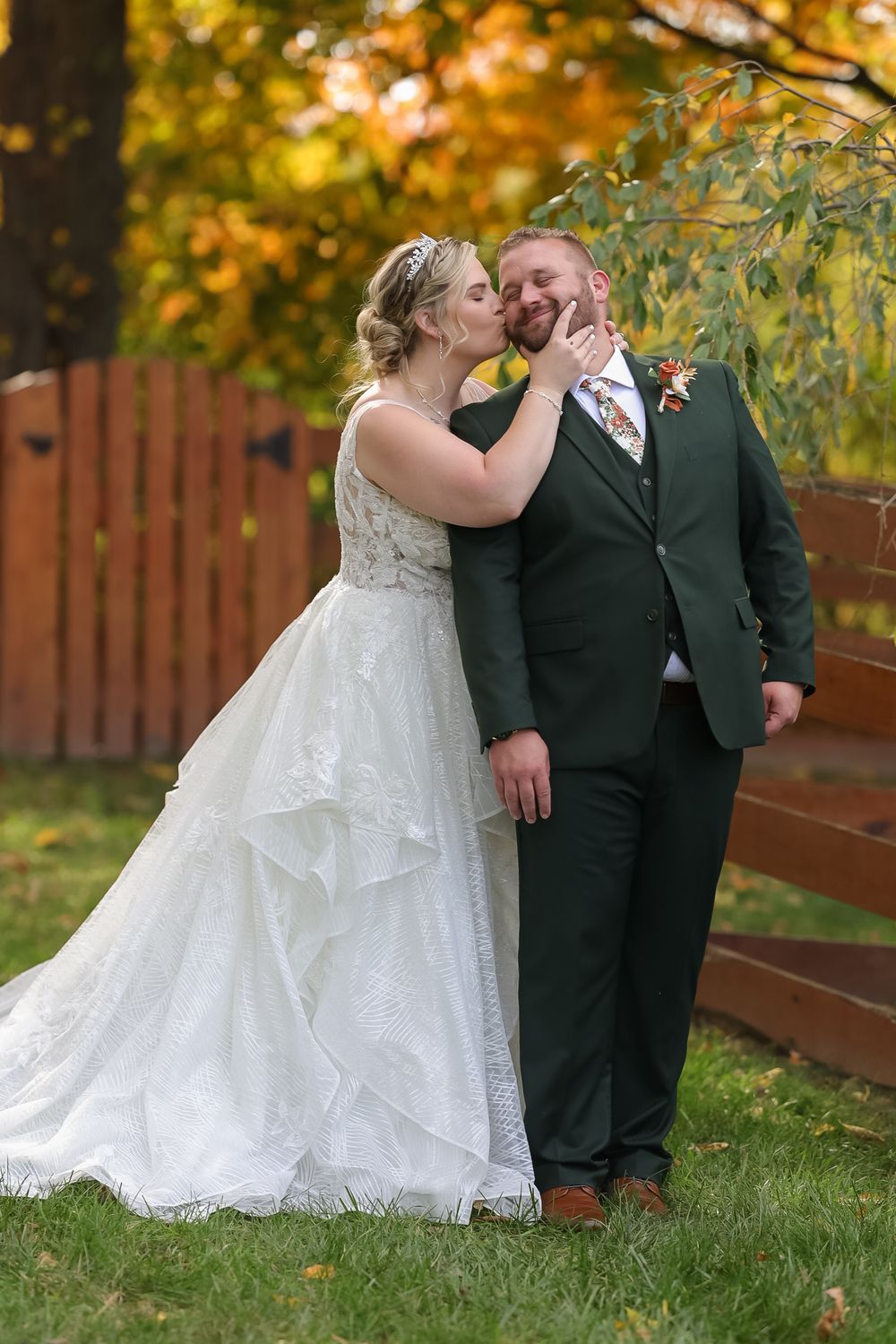 Bride kissing groom on the cheek while he smiles in a green suit against a fall foliage backdrop.