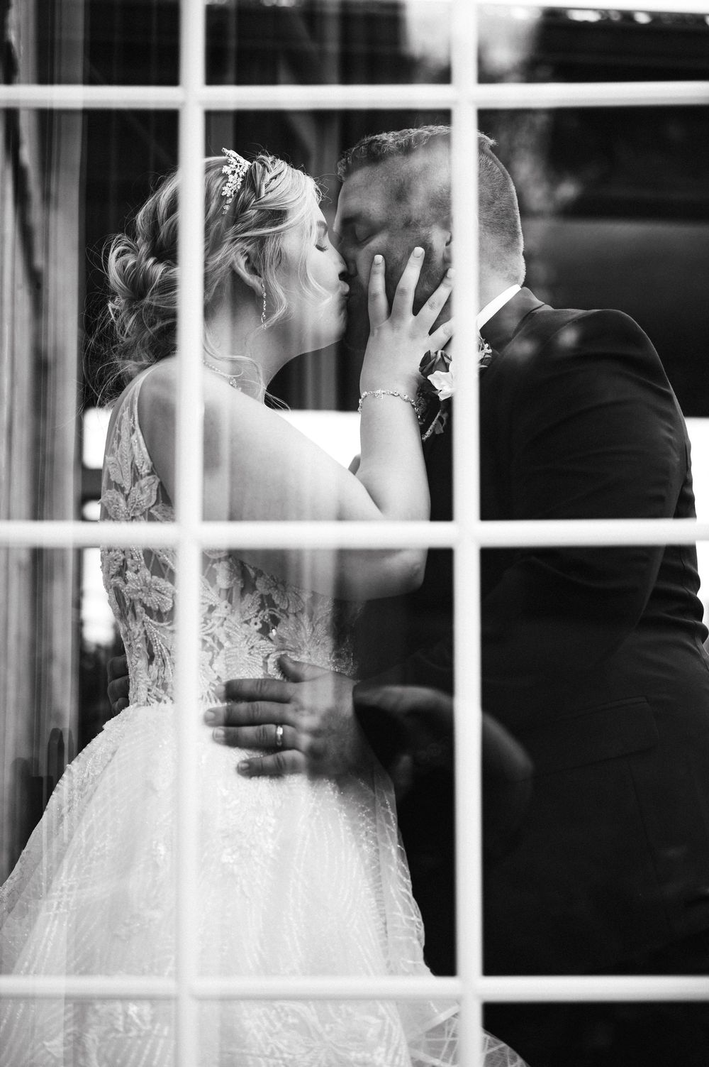 Black and white photo of bride and groom kissing passionately reflected through a large window pane.