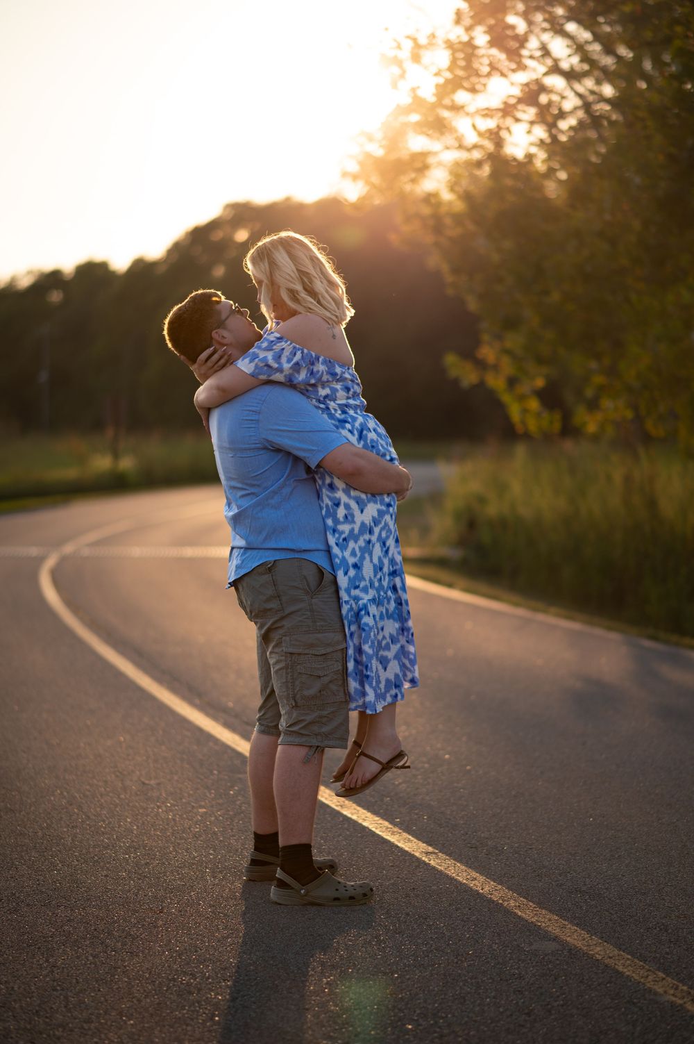 Couple embracing on a sunlit road at golden hour with trees in the background.