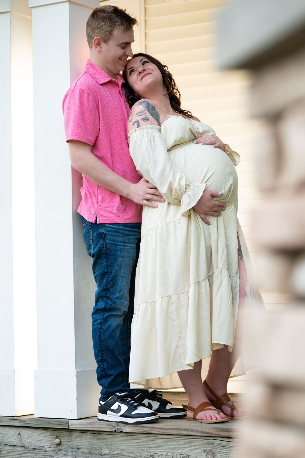 Couple embracing on porch, pregnant woman in cream dress with partner in pink shirt and jeans.