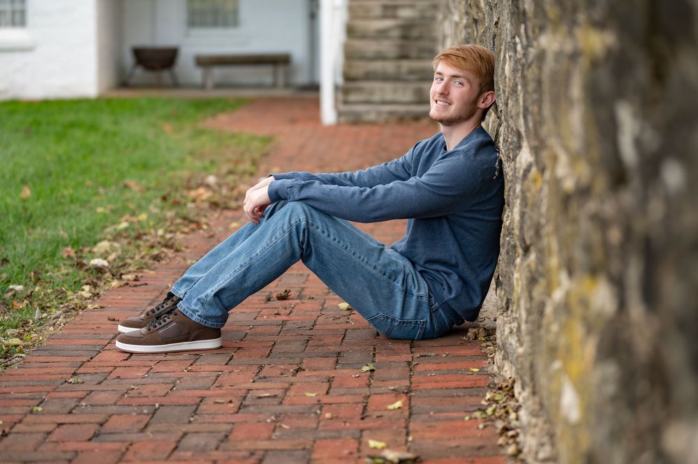 Young red-haired man sitting against brick wall on red brick pathway outdoors.