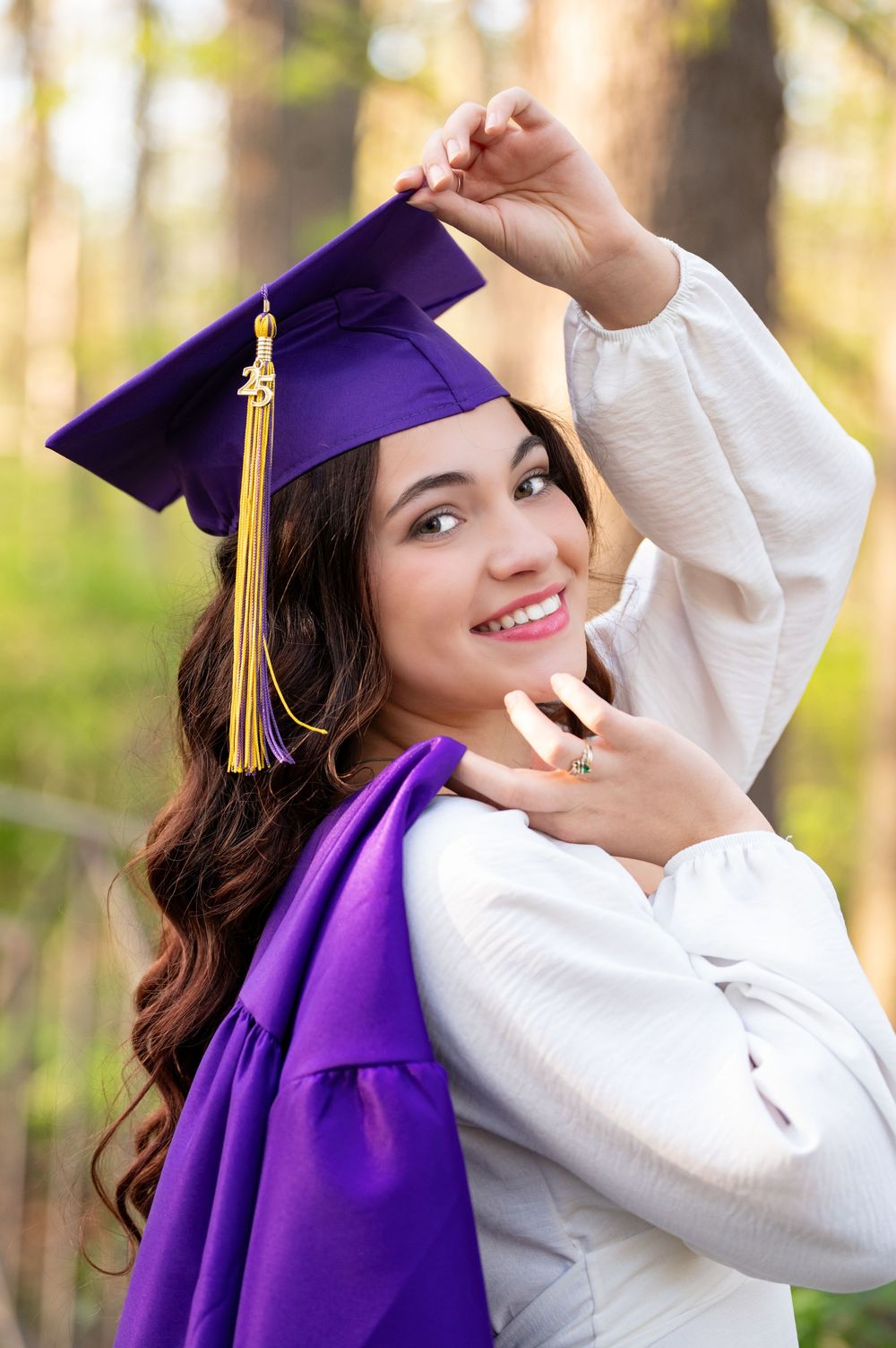 Smiling young woman in purple graduation cap and gown posing outdoors among trees.