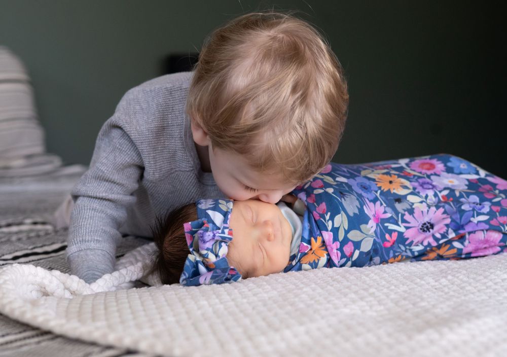 Toddler boy gently kissing swaddled newborn baby in floral blanket on white surface.