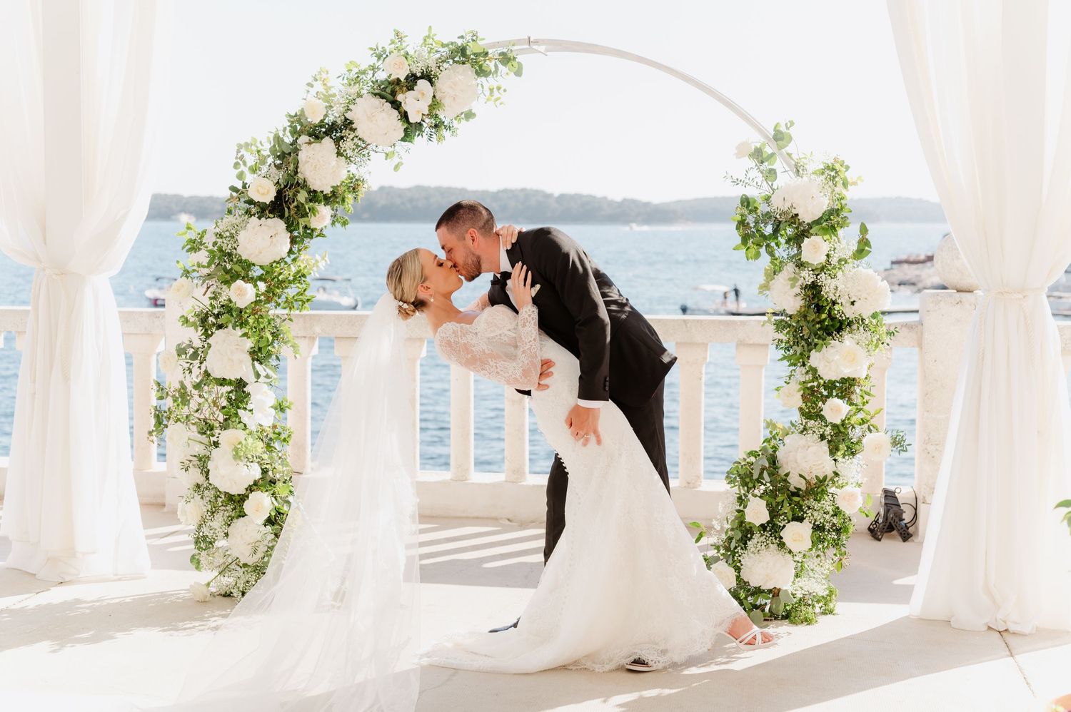Romantic wedding moment under a white floral arch overlooking the ocean.