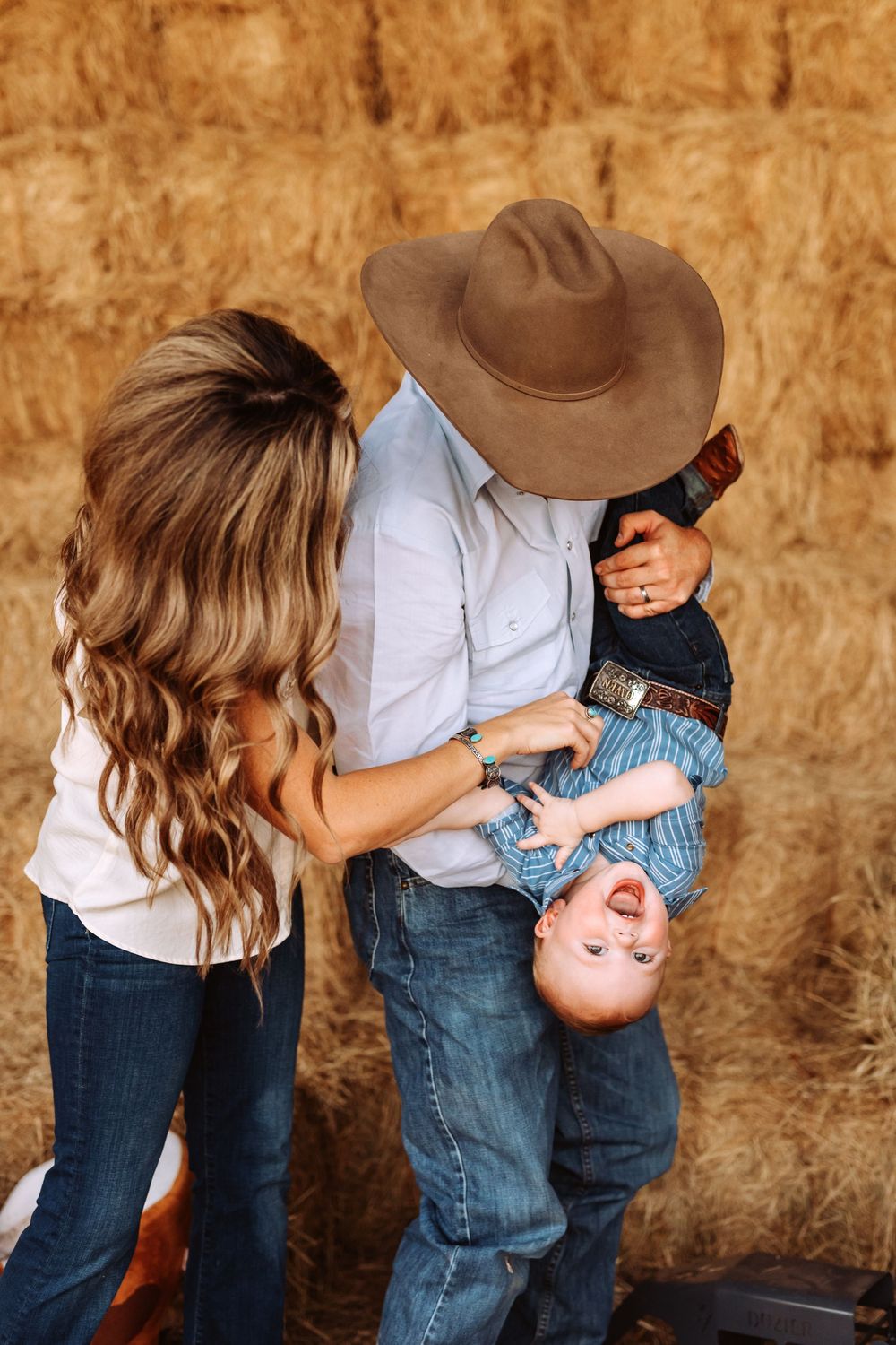 mom and dad playing with toddler boy. people in jeans and white shirts against a rustic barn wall background. San Marcos family photographer