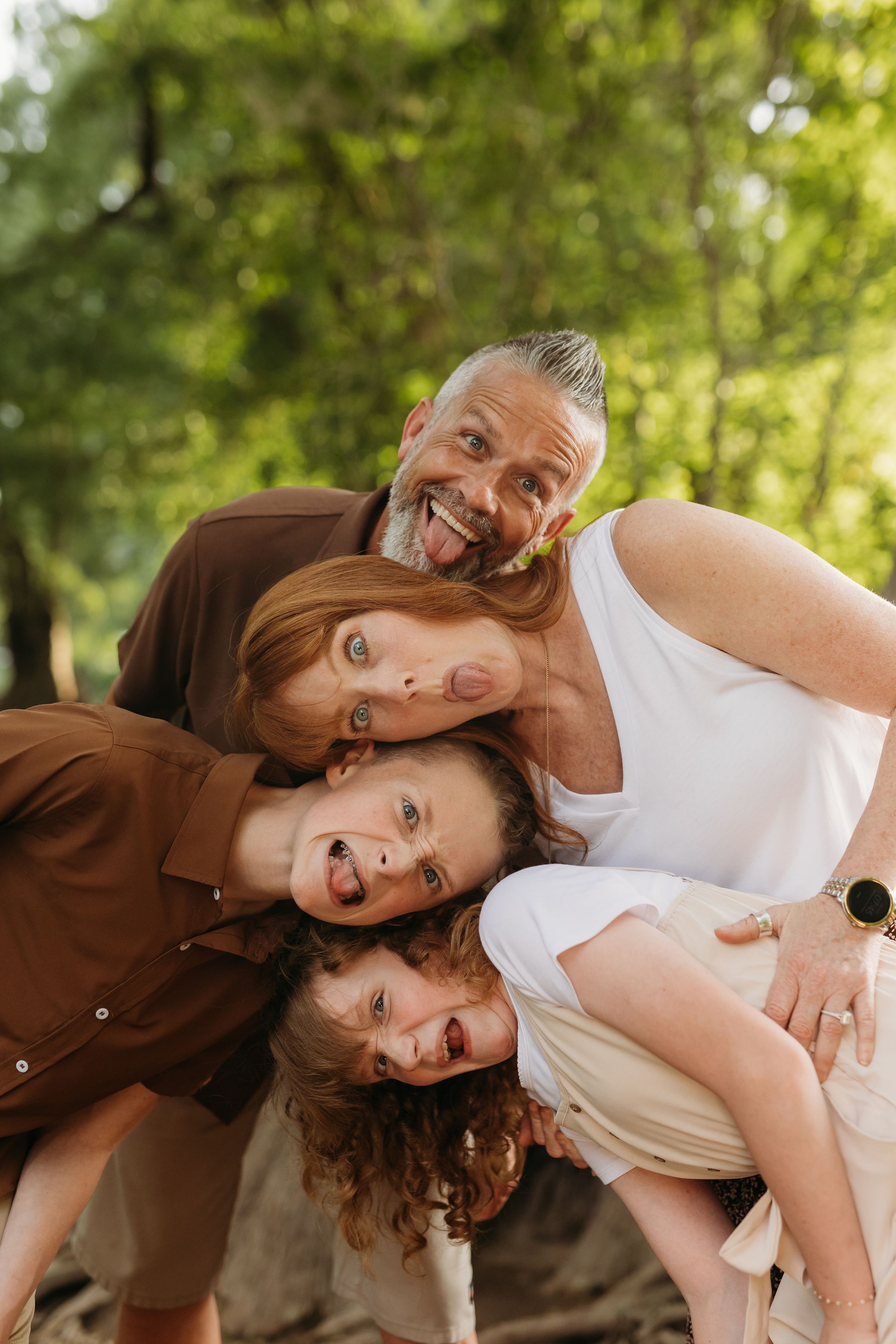 A joyful family of four making silly faces together outdoors in a lush green park setting. San Marcos family photographer