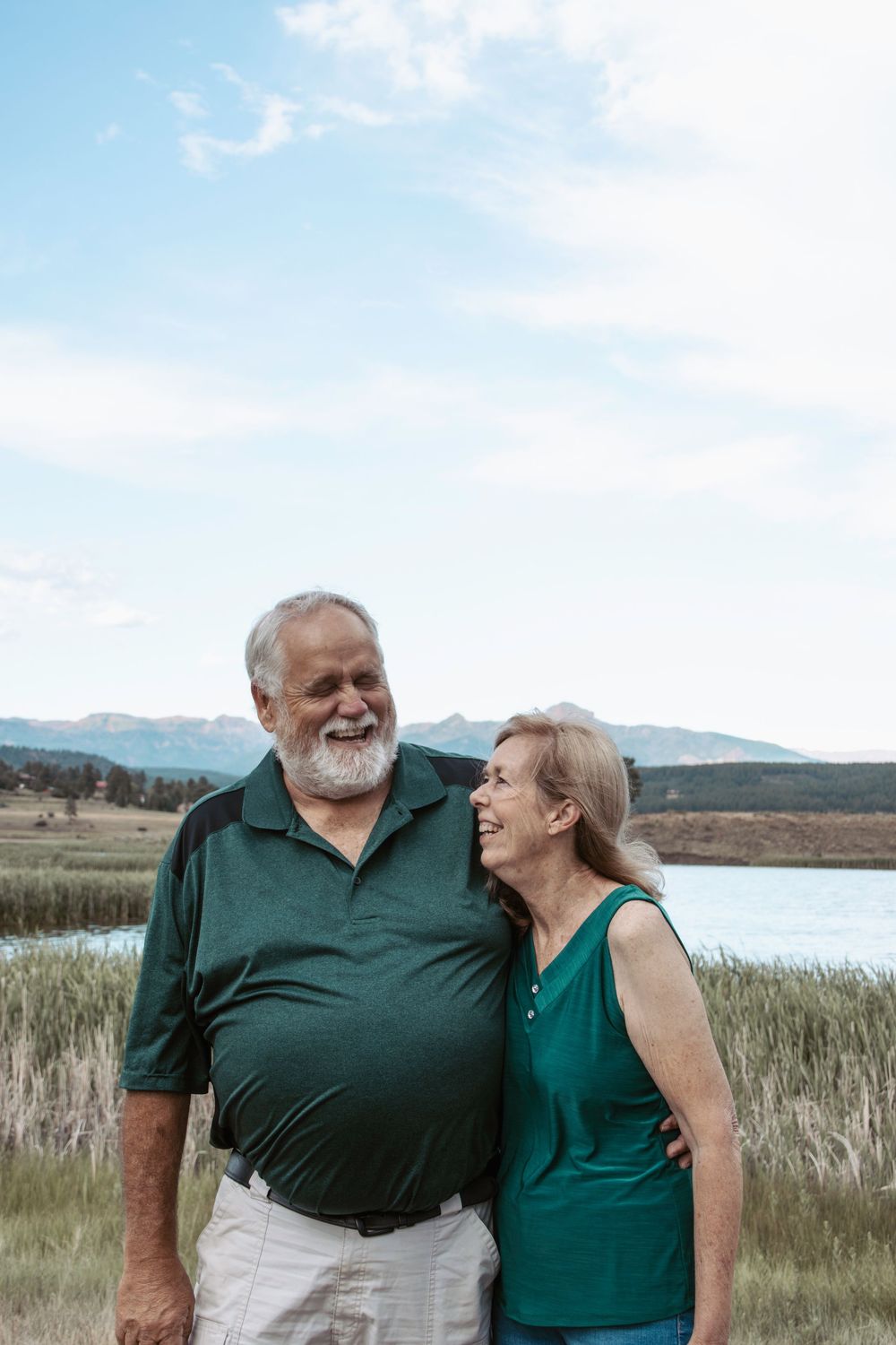 A couple in green shirts share a tender moment together while standing in front of a scenic lake and grassy landscape.