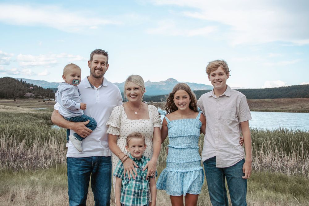 A group stands together smiling in front of a scenic mountain lake landscape on a sunny day.